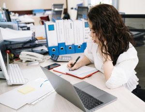 Woman at desk surrounded by binders, papers, computers, and a calculator, completing the annual nonprofit audit process, with a look of frustration on her face.