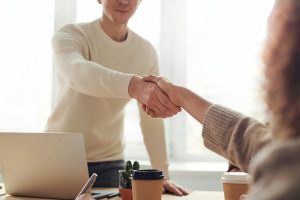 Man reaches out to shake a woman's hand over a desk that is covered in laptops and paperwork, as if to extend a job offer.