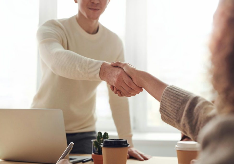 Man reaches out to shake a woman's hand over a desk that is covered in laptops and paperwork, as if to extend a job offer.